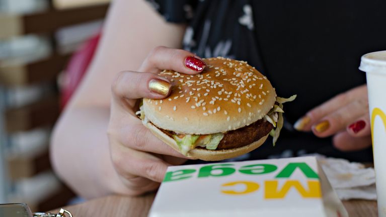 Nainen, jolla on kynsissään punaista ja kultaista kynsilakkaa, syö McVegan -hampurilaista.