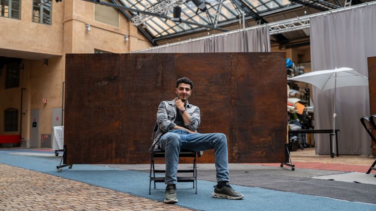 Näyttelijä Yussef Asab Alkhatib istuu rennosti Teatterikorkeakoulun avoimella sisäpihalla ruosteinen lavastetausta takanaan.