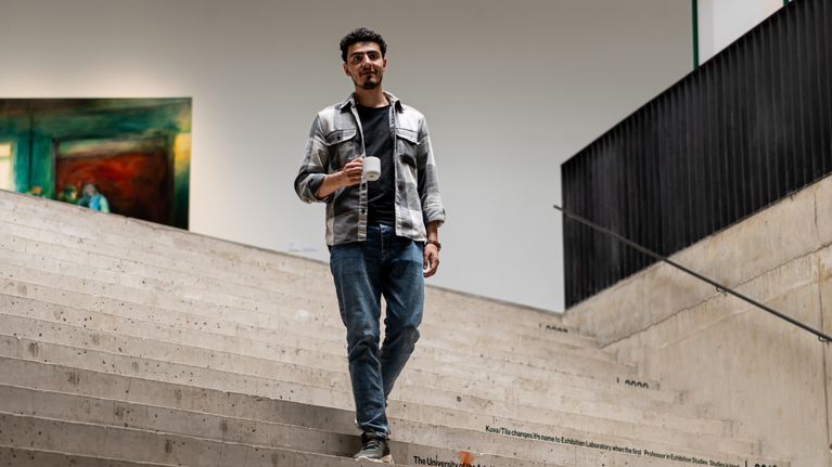 Näyttelijä Yussef Asab Alkhatib kävelee kahvikuppi kädessä alas puhtaita betonisia sisäportaita.