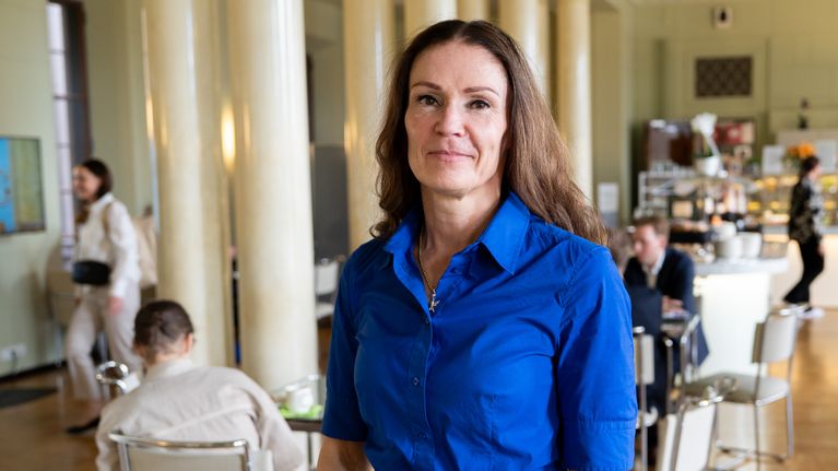 Lulu Ranne står i riksdagens kafé i en blå klänning och tittar mot kameran.