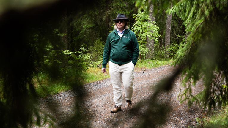 Henkilö kävelee metsäpolulla havupuiden katveessa, taustalla näkyy metsää ja sorainen polku.
