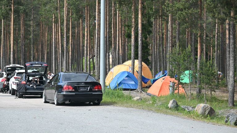 Autoja pysäköitynä soratien varrelle metsän reunaan, taustalla useita värikkäitä telttoja puiden keskellä.