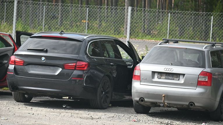 Parkkipaikalla on kolme autoa ilman kilpiä, joista kahden takaovet ovat auki. Taustalla näkyy metalliaita ja metsää.