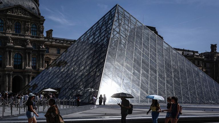 Ihmisiä kävelee ja seisoo lasisen pyramidin edessä Louvren museon aukiolla aurinkoisena päivänä.