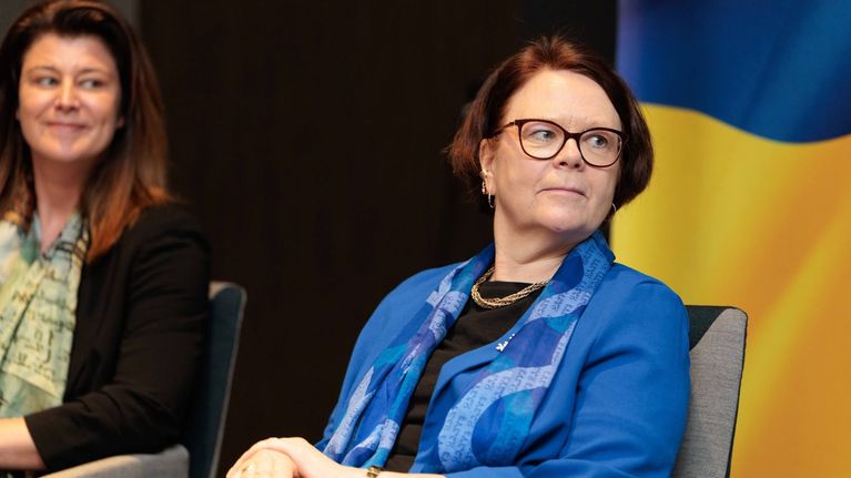 Nainen sinisessä bleiserissä istuu tuolilla ja katsoo ohi kameran.