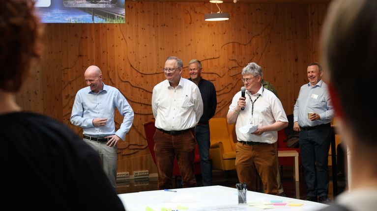iisi miestä seisoo rivissä sisätiloissa, yhdellä on mikrofoni kädessään. Yläkulmassa on suuri valokuvaprojektio.