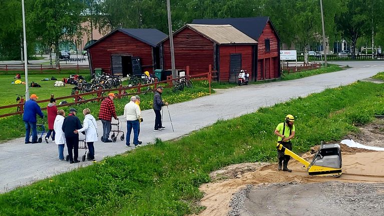 Ryhmä ihmisiä kävelee soratiellä, taustalla punaisia puurakennuksia ja edustalla työmaamies käyttää tärylevyä.