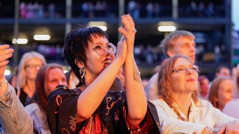 Ihmisiä seisoo ulkona konsertissa iloisina katsomassa lavalle ja taputtamassa käsiään.