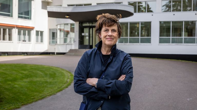 Mirkku Kullberg seisoo kädet ristissä parantolarakennuksen edessä asfaltoidulla pihalla.