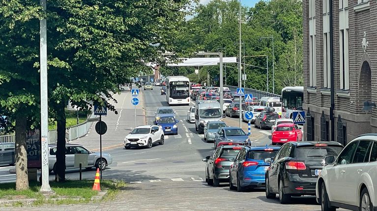 Ruuhka kaupungin keskustan kadulla kesäisenä päivänä. Vasemmalla lehtipuu, oikealla tiiliseinäinen rakennus.