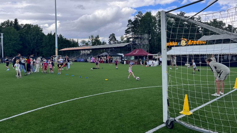 Lapset pelaavat jalkapalloa nurmikentällä, taustalla katoksia ja ihmisiä sekä rakennuksessa teksti “Areena”.