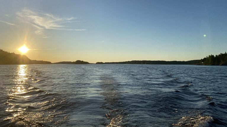Veneen peräaallot järvellä ilta-auringossa, taustalla näkyy metsäistä rantaa ja saaria.