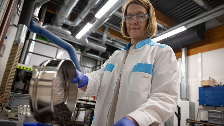 Polkkahiuksinen ja silmälasipäinen nainen kaataa metallisesta kulhosta tummia pellettejä astiaan. Päällä valkoinen labratakki.
