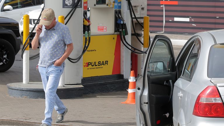 Mies kävelee kännykkä korvalla bensapumpun edessä. Vieressä on auto, jonka ovi on auki.