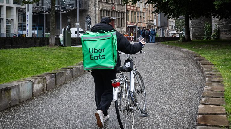 Henkilö taluttaa polkupyörää ja kantaa vihreää Uber Eats -reppua katuympäristössä, taustalla useita ihmisiä.