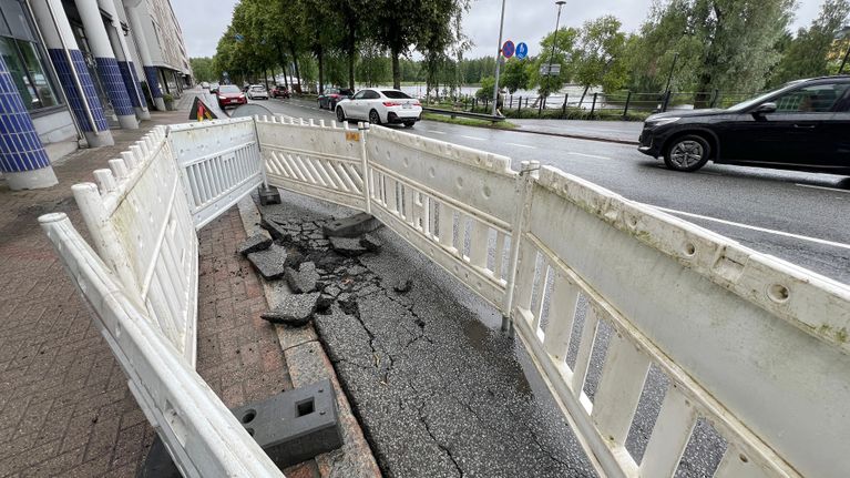 Asfaltti rikkoutunut kadun reunasta jalkakäytävän vierestä. Rikkoutunut alue ympäröity valkoisella työmaa-aidalla, taustalla autoja kadulla.