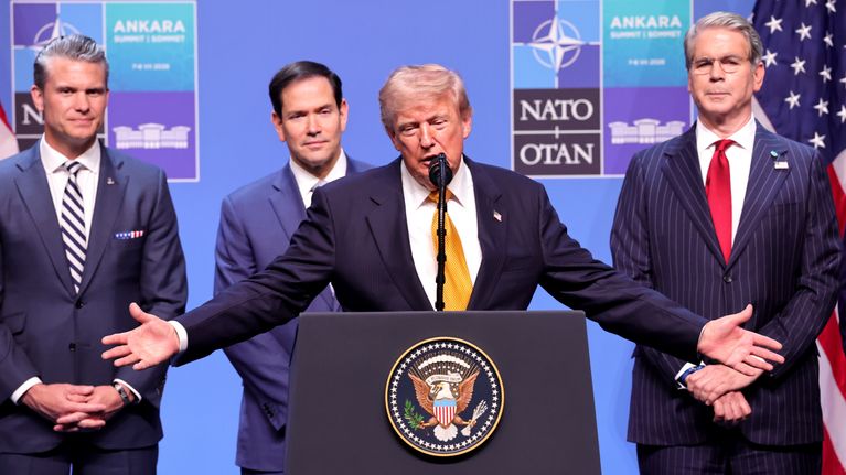 Donald Trump keltaisessa kravatissa puhuu puhujakorokkeella, taustalla Naton logo.