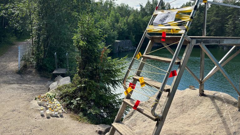 Uimahypputorni on teipattu keltaisella ja punaisella teipillä, portaissa on varoituslappu. Taustalla vettä ja metsää.