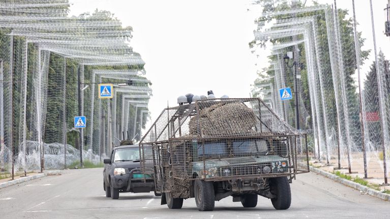 Drooniverkolla katetulla tiellä ajaa sotilasajoneuvo, joka sekin on varustettu suojaverkoilla. Perässä tulee toinen auto.