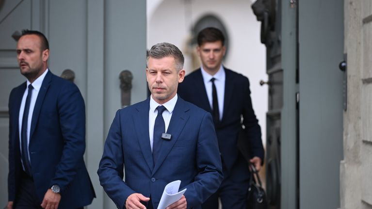 Vakava tummansiniseen pukuun pukeutunut keski-ikäinen mies kävelee paperipino kädessään ulos vanhoista puuovista, taustalla muita pukumiehiä.