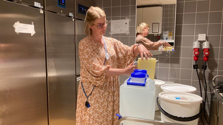 Nainen laittaa vesikannuun kantta. Pöydällä on kolme kannellista sankoa ja kaksi hanallista vesisäiliötä.