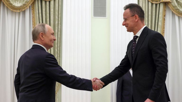 Putin ja Szijjártó kättelevät vaaleassa salissa. Heillä on tummat puvut, Szijjártólla on pilkullinen kravatti ja silmälasit.