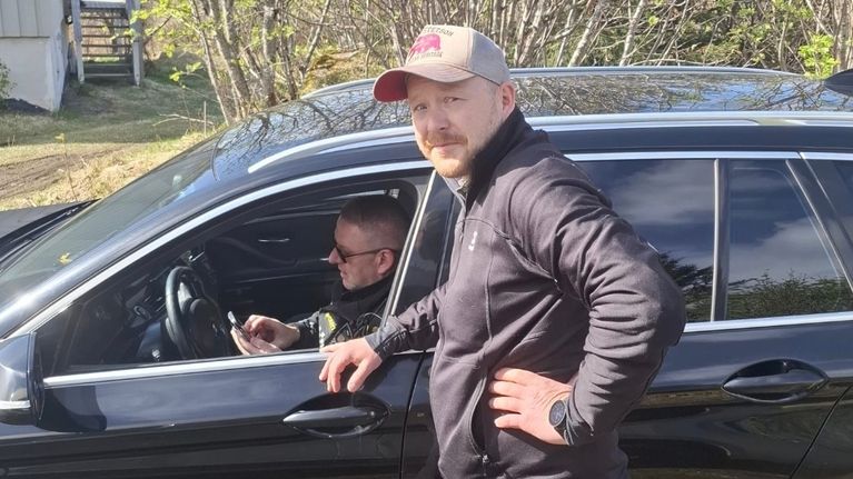 Poliisi istuu mustassa autossa ja kirjoittaa sakkoja. Autoon nojailee mies, joka katsoo kameraa hämmentyneen näköisenä.