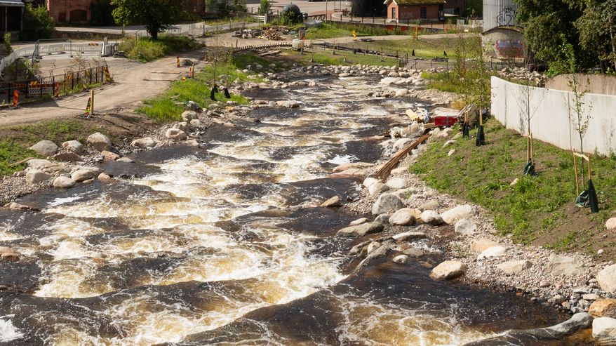 Tourujoki virtaa kuohuen. Taustalla näkyy asuikerrostaloja ja vanha tiilipiippu.