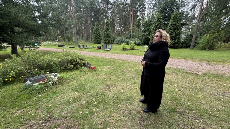 Nainen seisoo mustissa vaatteissa hautausmaalla. Hänen edessään on kaadettu hautakivi.