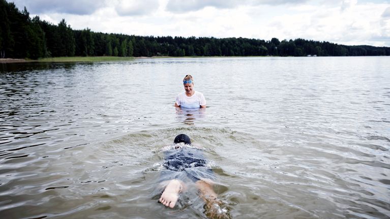 Kaksi henkilöä on järvivedessä. Toinen ui vedessä ja toinen seisoo vedessä katsellen häntä.