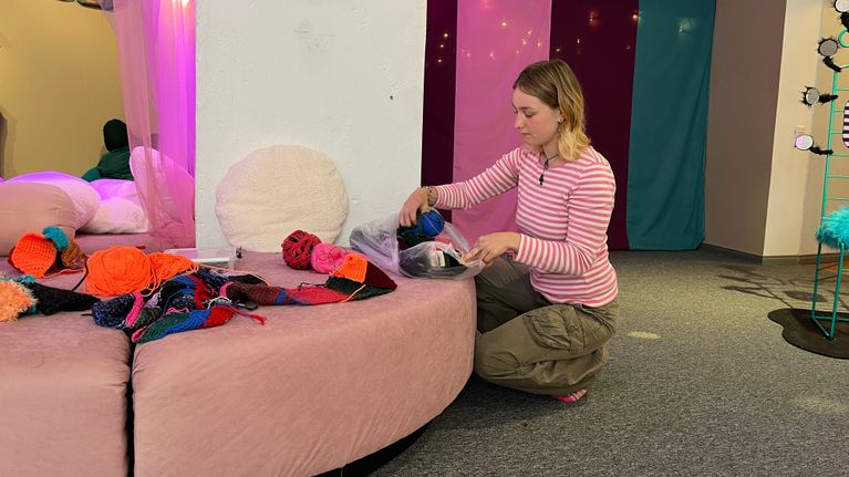 Nainen on kyykyssä vaaleanpunaisen sohvan vieressä ja kaivaa villalankakeriä muovipussista.