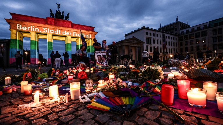 Brandenburgin portti on valaistu sateenkaaren väreissä, edustalla kukkia, kynttilöitä, sateenkaarilippuja ja valokuvia.