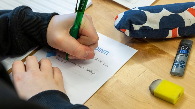 Vasenkätinen kirjoittaja kirjoittaa lyijykynällä paperille tekstaten. Pöydällä on myös kumi ja penaali.