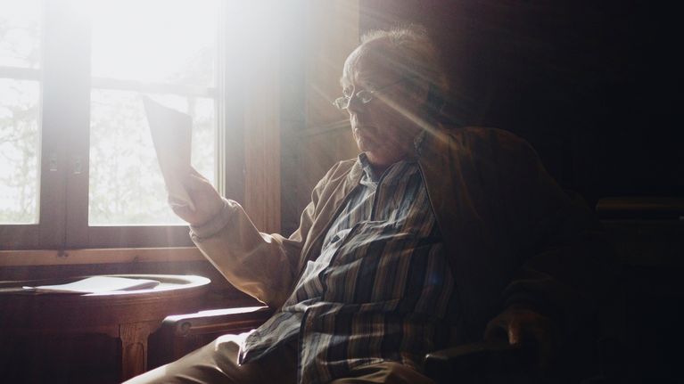 Mies istuu tuolilla ja pitelee paperia kädessään, kun aurinko paistaa kirkkaana ikkunasta.
