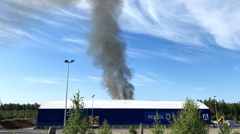 Matalan teollisuushallin takaa nousee musta savupatsas taivaalle.