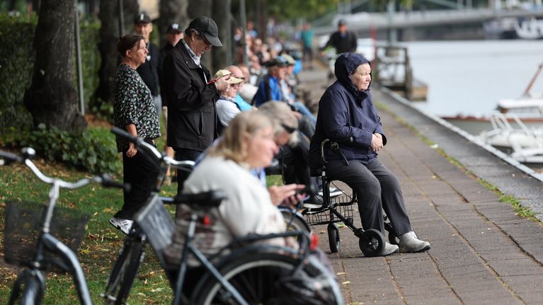 Ihmisiä seisoskelemassa ja istumassa Aurajoen rannalla ja katselemassa joen toiselle puolelle.