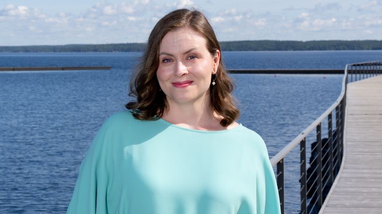 Nainen hymyilee järven rannalla.