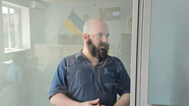 Parrakas Roope Mustonen seisoo syytetyn lasisessa kopissa oikeussalissa sinisessä paidassa. Kopin lasiin heijastuu tuomarin pöytä ja Ukrainan lippu.