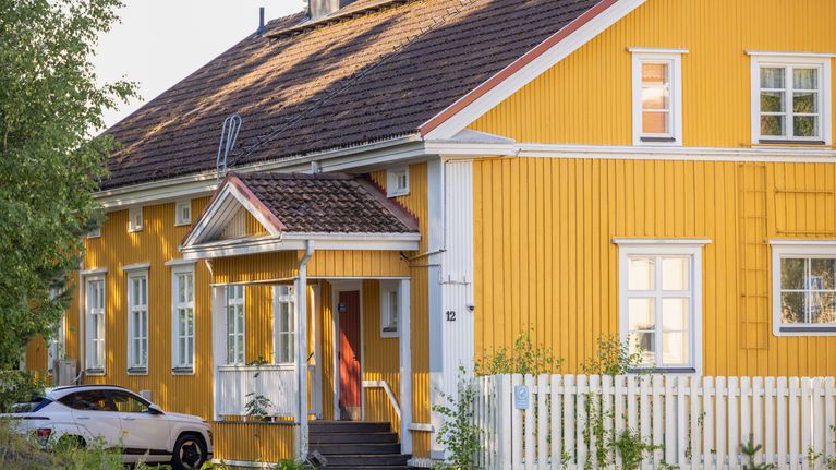 Keltainen puutalo kuvattuna aamuauringossa. Takapihalla on pysäköitynä auto.