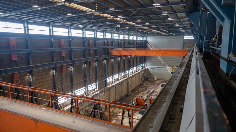 Teollisuushalli, jossa suuri nosturi ja keskeneräinen betoniallas; seinillä oransseja laitteita.