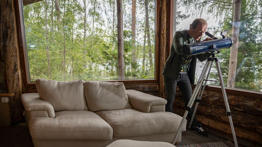 Henkilö katsoo kaukoputkella suuren ikkunan läpi metsäiseen maisemaan, vieressä vaalea sohva.