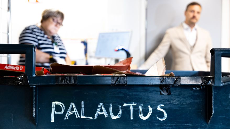 Etualallaroskalaatikko, jossa teksti "PALAUTUS"; taustalla pöydän ääressä istuva henkilö ja seisova henkilö.