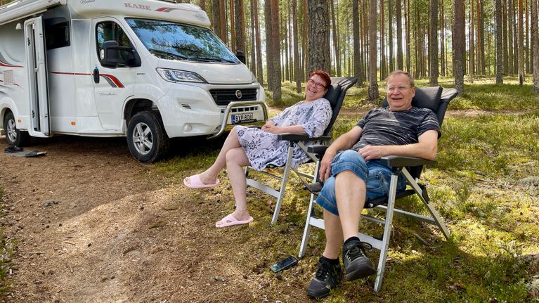Valkoinen matkailuauto parkissa mäntykankaalla ja sen edustalla hymyilevä pariskunta Sirpa ja Hemi Hyvvönen istuvat retkituoleissa.