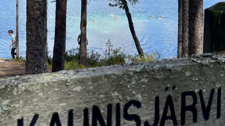 Kaunisjärven kyltti rannalla a takana vedessä uimareita.
