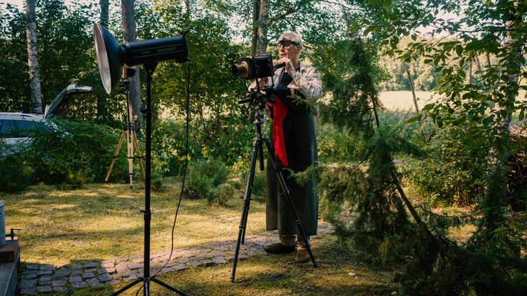 Henkilö seisoo ulkona valokuvauskaluston, suuren kameran ja salamalaitteen, vieressä metsäisessä ympäristössä.
