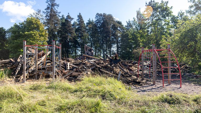 Leikkikentän kiipeilytelineet punaisine metallirakenteineen ja taustalla rakennuksen palanut puurunko sekä metsää.