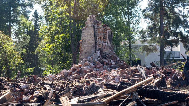 Tiilimurskaa ja raunioitunut savupiippu puiden keskellä, taustalla rakennuksia.