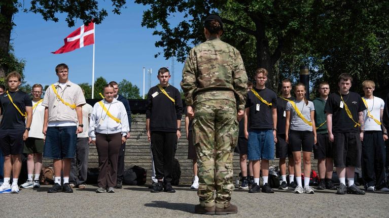 Maastopukuun pukeutunut nainen seisoo selin kameraan, taustalla rivissä seisoo nuoria aikuisia siviilivaatteissa.