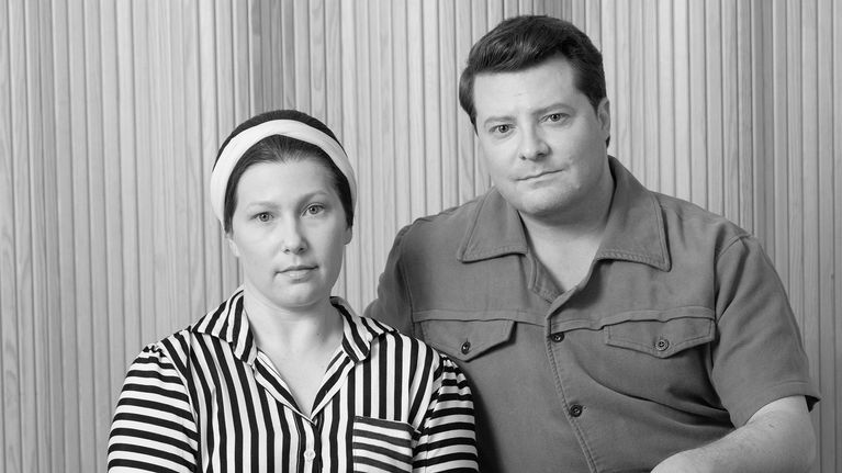 Teatteriesityksen kuva, nainen vasemmalla, mies oikealla istuvat ja katsovat suoraan kameraan. Vaatteet hieman 1950-lukua.