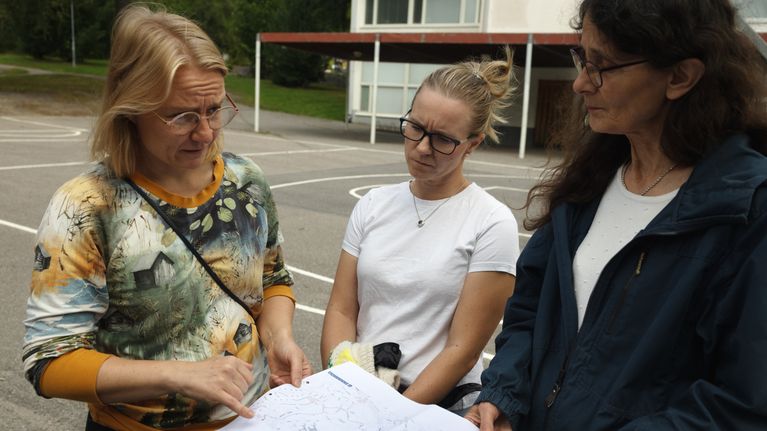 Kolme naista seisoo ulkona ja tutkii paperia, jossa on karttoja ja tekstiä. Taustalla näkyy rakennus ja piha-aluetta.